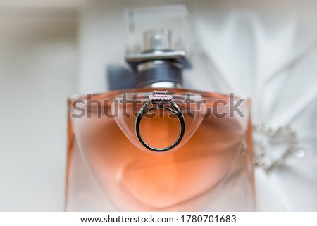 Top view picture of white gold diamond engagement ring placed on the bottle of peach color perfume, white wedding dress on the background