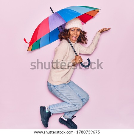 Young beautiful latin woman holding umbrella smiling happy. Jumping with smile on face over isolated pink background