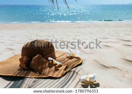 Straw hat with frangipani flowers on white sea sand. With beautiful seascape, the blue sea in the background,Palm leaf shadow. Copy space, background for advertisement of a seaside vacation