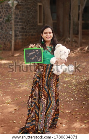 Pregnant Indian lady with teddy and baby coming soon board in hand.