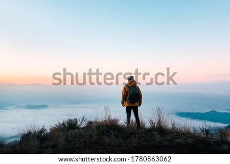 Back person contemplating an immense landscape covered with fog from the top of a mountain at sunrise. Traveller escaping in solitude into the wilderness. Connection to nature to the great outdoors.