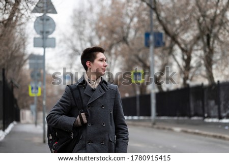 A young free handsome fashion guy dressed in a coat goes out with a backpack from home to the street to go on a trip or to study, urban style