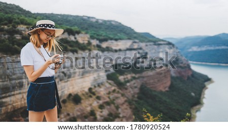young blonde girl takes photo of  panorama landscape on retro camera during  trip on mountains outdoors, hipster tourist in summer hat enjoys hobby of photographing nature spain on weekend empty space