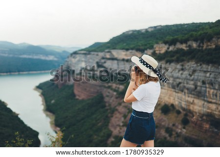 young blonde girl takes photo of  landscape on retro camera during  trip on mountains, hipster tourist in summer hat enjoys hobby of photographing beautiful nature on weekend empty space