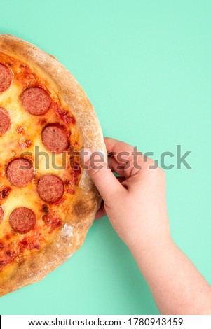 Vertical image with a woman's hand grabbing a pepperoni pizza. Top view image with a freshly baked pepperoni pizza on a green-mint background.