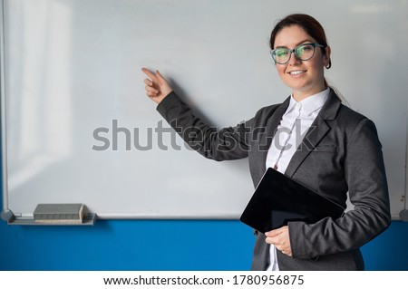Beautiful woman in a suit with a digital tablet in her hands. A smiling female business coach with glasses stands in a conference room and points a finger at a white board. Workshop at the office. Royalty-Free Stock Photo #1780956875