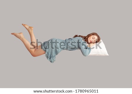 Relaxed girl in vintage ruffle dress levitating in mid-air, sleeping on stomach lying comfortable cozy on pillow, keeping eyes closed, watching peaceful dream. indoor studio shot isolated on gray Royalty-Free Stock Photo #1780965011