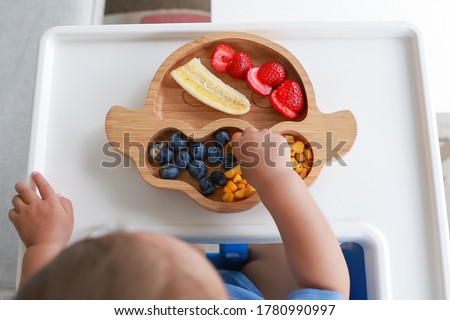 Top view Baby self-feeding with hand BLW or baby led weaning. Finger food plate of mix fruit strawberry, banana, blueberry and corn. Kid healthy nutrition eating on high chair fine motor development. Royalty-Free Stock Photo #1780990997