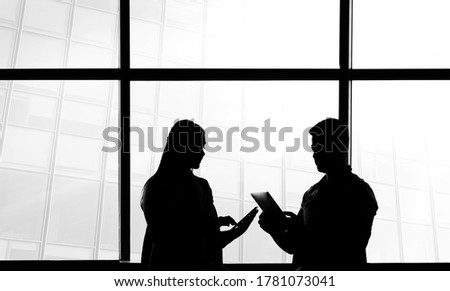 Silhouettes of 2 two business people man and woman sitting on armchairs in office interior, man showing information on screen of his tablet to his colleague, huge windows with cityscape outside