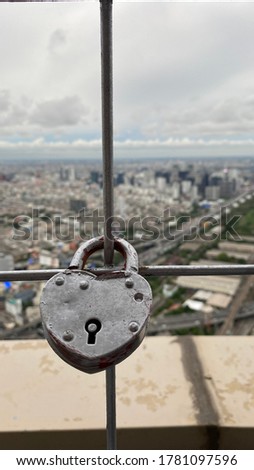 A photo of a heart shaped lock locked on a fence of a high building with Bangkok city blurry view on the background. 