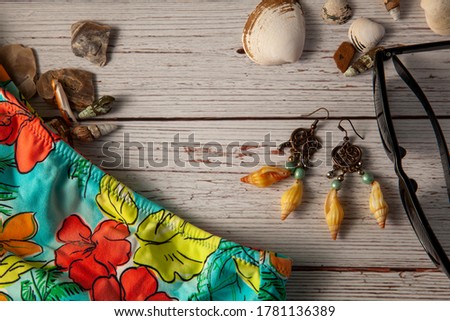A flat lay photography showing a composition of beach vacation items including a pair of sunglasses, a girl's swimsuit, sea shells, decorative stones, earrings on wooden background.