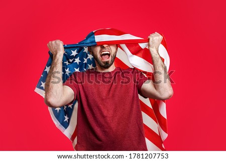 Caucasian man with an American flag on his head throws his hands up and shouts. Red background. The concept of patriotism, power, freedom and sports fans