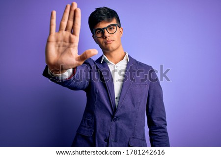 Young handsome business man wearing jacket and glasses over isolated purple background doing stop sing with palm of the hand. Warning expression with negative and serious gesture on the face.
