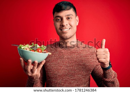Young hispanic man eating vegetarian healthy salad over red isolated background happy with big smile doing ok sign, thumb up with fingers, excellent sign