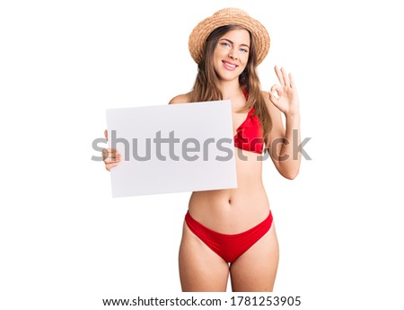 Beautiful caucasian young woman wearing bikini holding blank empty banner doing ok sign with fingers, smiling friendly gesturing excellent symbol 