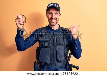 Handsome policeman wearing uniform and bulletprof holding handcuffs over yellow background pointing thumb up to the side smiling happy with open mouth