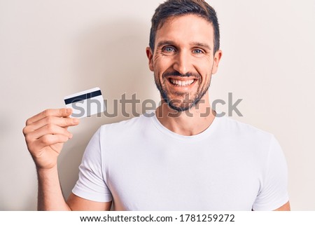 Young handsome customer man holding credit card over isolated white background looking positive and happy standing and smiling with a confident smile showing teeth