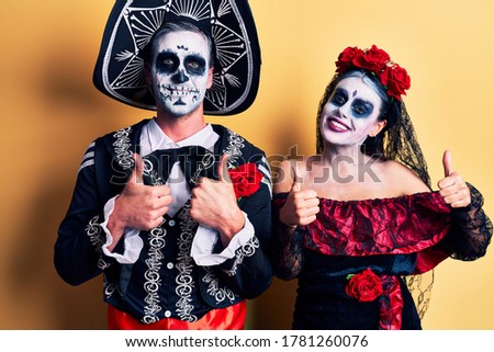 Young couple wearing mexican day of the dead costume over yellow success sign doing positive gesture with hand, thumbs up smiling and happy. cheerful expression and winner gesture. 