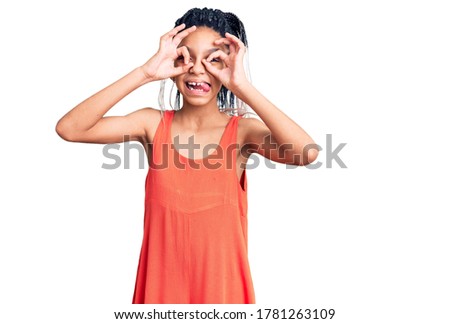 Cute african american girl wearing casual clothes doing ok gesture like binoculars sticking tongue out, eyes looking through fingers. crazy expression. 