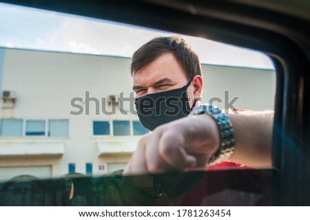 A man in a black mask and a red T-shirt stands leaning on a car window in a warehouse