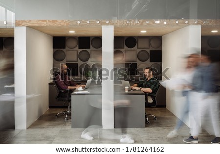 Two diverse businessmen working at desks in cubicles with blurred colleagues walking around a busy office