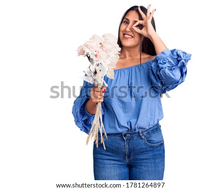 Young beautiful brunette woman holding bouquet smiling happy doing ok sign with hand on eye looking through fingers 