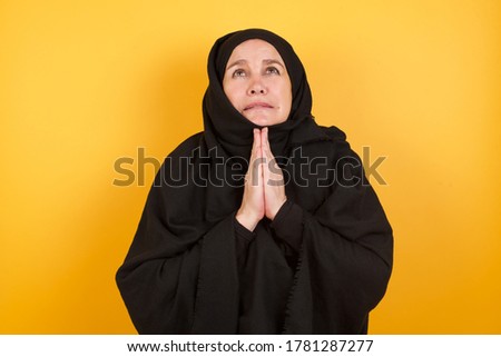 Beautiful middle aged muslim woman wearing black hijab over yellow background begging and praying with hands together with hope expression on face very emotional and worried. Asking