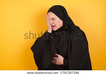 Beautiful middle aged muslim woman wearing black hijab over yellow background Yawning tired covering half face, eye and mouth with hand. Face hurts in pain.