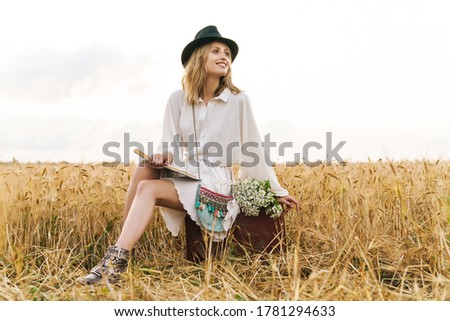 Image of young blond caucasian woman reading book while sitting in golden field on countryside