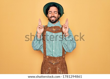 Young handsome man wearing tratidional german octoberfest custome for Germany festival gesturing finger crossed smiling with hope and eyes closed. Luck and superstitious concept.