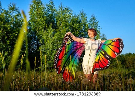 Young cute pretty woman with bright fabric like butterfly wings play, fly, run, jump in meadow or field with green grass. Girl or female dancer having fun on nature landscape during sunset