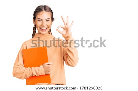 Beautiful caucasian woman with blonde hair holding book doing ok sign with fingers, smiling friendly gesturing excellent symbol 