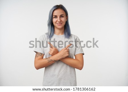 Charming young blue-eyed tattooed lady with short haircut looking positively at camera while pointing with forefingers in different directions, isolated over white background