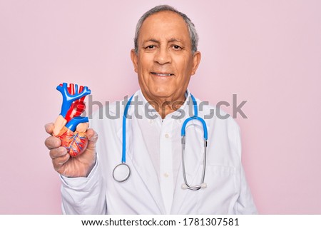 Senior hoary doctor man wearing stethoscope holding plastic heart over pink background with a happy face standing and smiling with a confident smile showing teeth