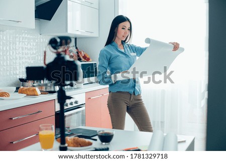 Beautiful calm woman standing in the kitchen and opening architectural paper roll. Camera on tripod on the table