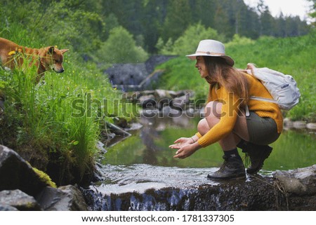 Happy young woman with dog standing by stream on a walk outdoors in summer nature.