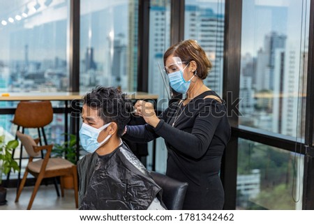 Young good looking male customer takes hair cut in a sky creeper business office while barber puts hygienic face mask and shield on to protect virus infection as a business in new normal. Royalty-Free Stock Photo #1781342462