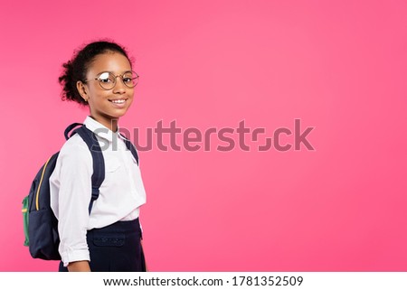 smiling african american schoolgirl in glasses with backpack isolated on pink