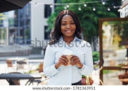 Holding cup of drink in hands. Young afro american woman in white shirt outdoors in the city near green trees and against business building.