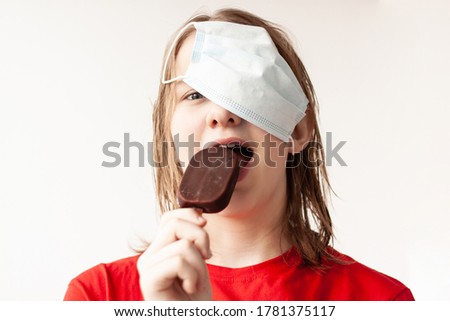 A teenage girl with blonde hair pushed a medical mask up on her hair, bites off a chocolate ice cream, selective focus.