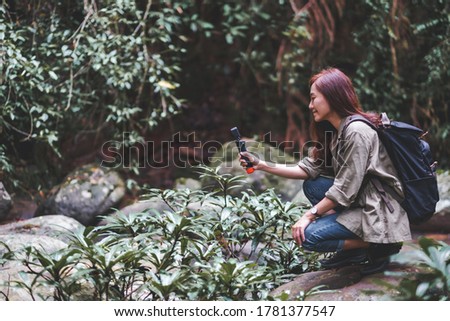 A female tourist with backpack shooting video of the waterfall in the forest by an action camera