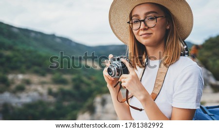 happy tourist women hold in hands retro camera trekking in nature outdoor, young blonde woman in hat and glasses leisure weekend in summer uses camera takes photos on background of mountain, concept