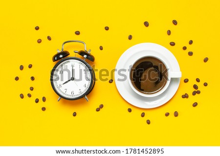 Cup of black coffee, coffee beans, black alarm clock on yellow background Flat lay top view copy space. Minimalistic food concept, morning breakfast, time to work, hot drink, coffee background