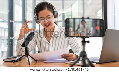 Asian women podcaster podcasting and recording online talk show at studio using headphones, professional microphone and computer laptop on table looking at camera for radio podcast.