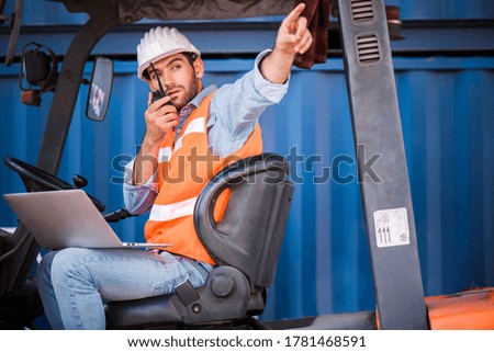 Young confident Caucasian man engineer driving Forklift with white safety helmet and check for control loading containers box by walkies talkie from Cargo freight ship for import and export, transport