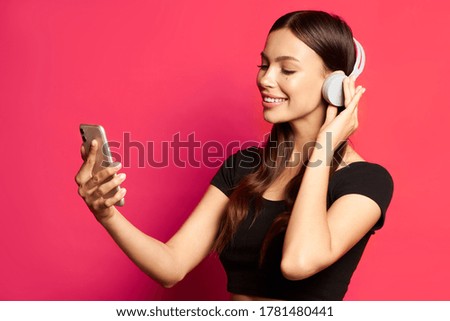 Girl listens to music and smiles in headphones. Choose music on the phone on a pink background.