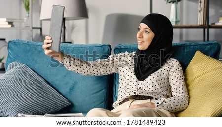 Portrait of a young happy muslim woman in hijab at home using digital tablet while sitting on sofa. 