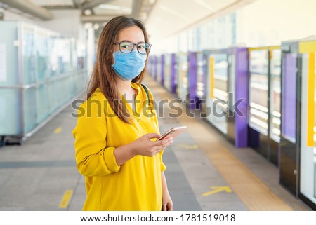 Young Asian business woman in casual shirt and medical mask working with her smartphone during covid-19 or corona virus at platform of Sky train station nearly office building. Bangkok, Thailand.