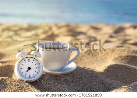 A white alarm clock and a cup of tea or coffee standing on the sand against the background of the blue sea at sunrise. Breakfast on the beach. Good morning concept