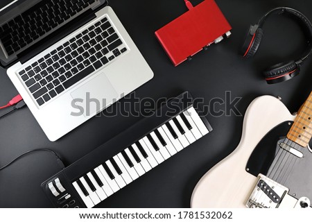 Top view of home recording studio equipment, electric guitar, MIDI keyboard, audio interface and headphone on black background.